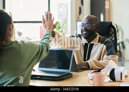 Young smiling male programmer giving high five to female colleague sitting in front of him during work over new software Stock Photo