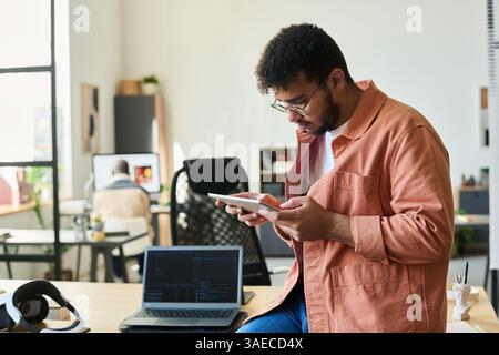 Young serious software developer with tablet working over new project while sitting on desk in coworking space Stock Photo