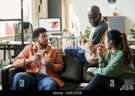 Two young male programmers and their female colleague having snacks and discussing new software at lunch break in office Stock Photo