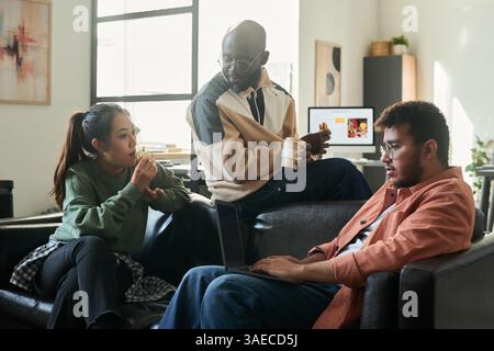 Young intercultural office workers sitting on leather sofa and discussing working points while having sandwiches and coffee Stock Photo