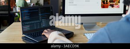 Header of two desktop computers and hand of young male programmer typing on laptop keypad while decoding data Stock Photo