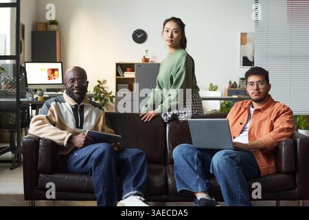 Team of young intercultural software developers with gadgets looking at camera while sitting on black leather sofa in office Stock Photo