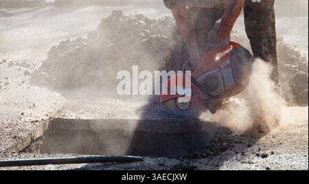 Construction worker using a power saw to cut through a tarmacked  driveway to access a bust pipe for repair on a sunny day Stock Photo