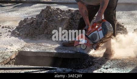 Construction worker using a power saw to cut through a tarmacked  driveway to access a bust pipe for repair on a sunny day Stock Photo