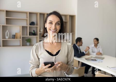 Smiling Latina businesswoman standing in conference room with smartphone Stock Photo