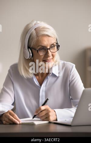 Interested older woman enjoy elearning with headphones on using notebook Stock Photo