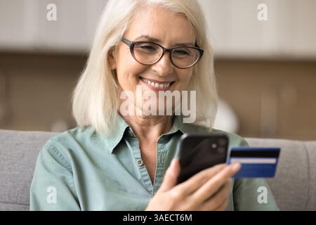 Happy aged lady sit on sofa hold cell credit card Stock Photo
