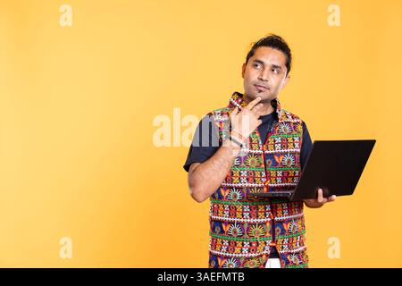 Pensive man debating how to solve business tasks on laptop, doing hand gesture, studio background. Employee does stroking chin gesturing during brainstorming process, using on notebook Stock Photo