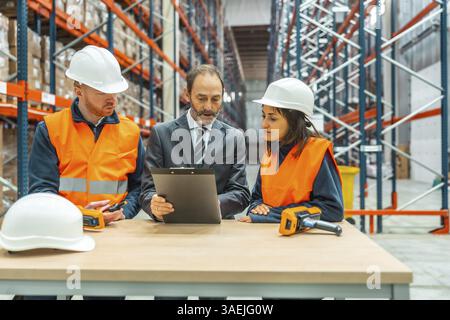 Warehouse manager presenting an inventory report on a clipboard while engaging with warehouse workers in a collaborative discussion Stock Photo