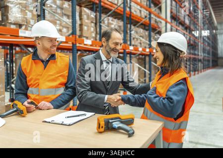 Manager shaking hands with a smiling female worker in a logistics warehouse, a male worker is looking at them, barcode readers and contract on the tab Stock Photo