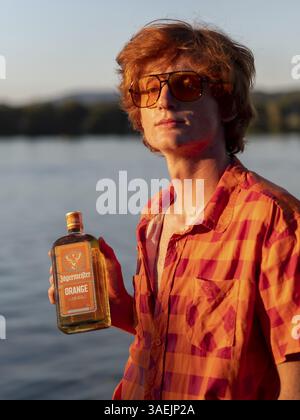 Young man standing by the lake, holding a bottle of alcohol in his hands, red hair, orange shirt, herbal liqueur, portrait Stock Photo