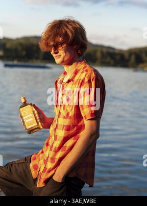 Young man standing by the lake, holding a bottle of alcohol in his hands, herbal liqueur, red hair, orange shirt Stock Photo