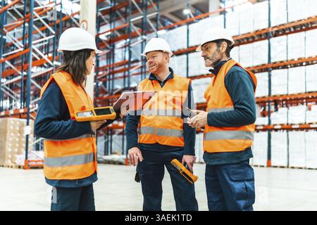 Warehouse workers wearing safety vests and helmets are using barcode readers and discussing logistics operations Stock Photo