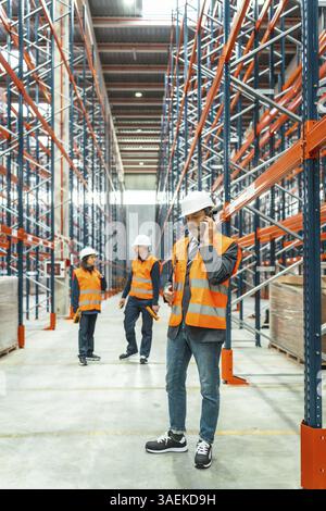 Warehouse manager wearing safety vest and helmet using smartphone, coordinating logistics operations with his team in large distribution center Stock Photo