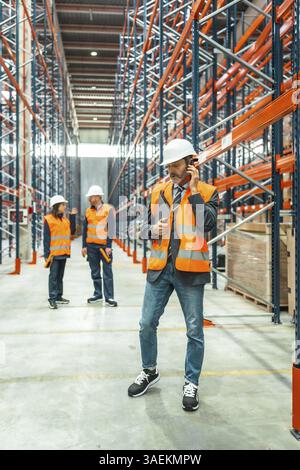 Manager coordinating logistics operations using phone and holding clipboard, workers wearing high visibility vests collaborating in warehouse environm Stock Photo