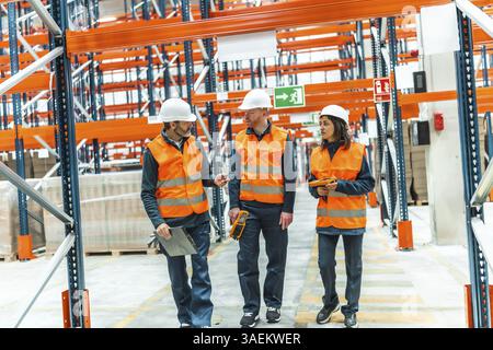 Warehouse workers wearing safety vests and helmets are walking through a large distribution center, discussing logistics and inventory management Stock Photo