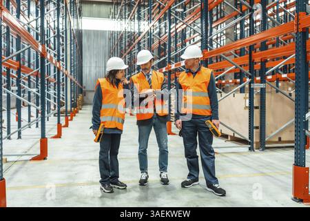 Warehouse workers and manager discussing logistics using a digital tablet, surrounded by shelves in a large distribution center Stock Photo