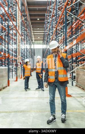 Warehouse manager wearing safety vest and helmet using mobile phone, coordinating logistics operations with his team in large distribution center Stock Photo