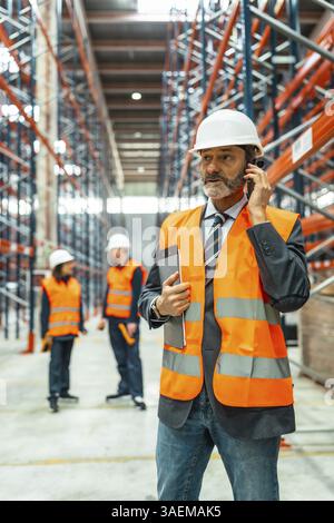 Warehouse manager wearing safety vest and helmet using phone and holding tablet while coordinating logistics operations Stock Photo