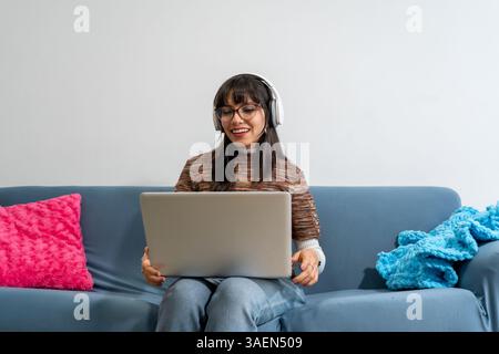 Happy young woman working remotely using laptop and headphones while sitting on a comfortable sofa Stock Photo