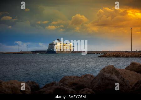 A cruise ship docked in a harbor under a dramatic, cloudy sky at dusk as a breakwater rocks in the foreground in Palma de Mallorca, Spain. Stock Photo