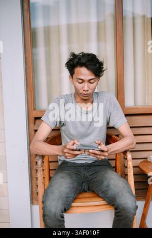 Young man absorbed in a phone game, relaxing in a chair on the veranda with his mobile device Stock Photo