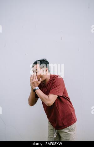 Worried man covering his face with hands standing against a white wall. Experiencing anxiety and feeling distressed. Stock Photo