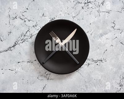 Black empty plate with crossed fork and knife on gray marble table, representing minimalist dining concept. Weight loss and dieting concept. Stock Photo