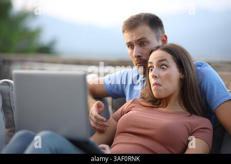 Confused couple watching media content on laptop sitting in a terrace at home Stock Photo