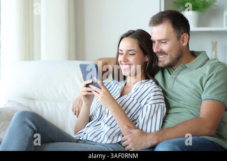 Happy couple reading together an ebook sitting on a couch at home Stock Photo