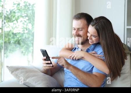 Happy couple using cell phone sitting in a couch in a house interior Stock Photo