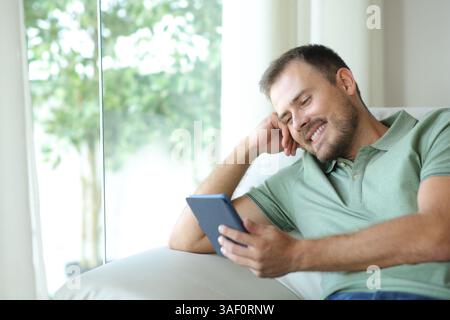 Happy man enjoying reading an ebook sitting on a sofa in a house interior Stock Photo