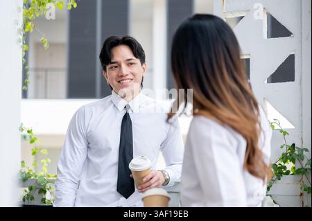 Asian male office worker is smiling at his female coworker while chatting together during a coffee break. Businessman and woman spending free time in Stock Photo