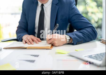 Close up of a man hand in blue suit holding a pen writing or noting down in a notebook. Working or having a meeting at office. Stock Photo