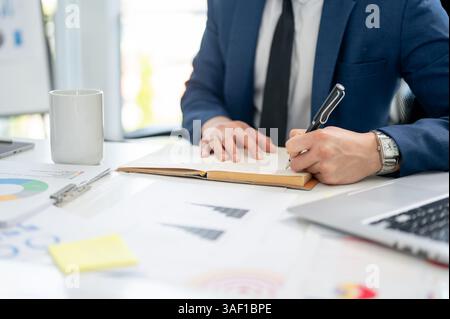 Close up of a man hand in blue suit holding a pen writing or noting down on in a notebook. Working or having a meeting at office. Stock Photo
