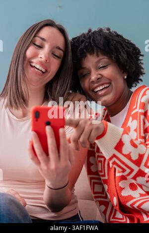 Two happy women sitting on couch smiling while looking at smartphone screen Stock Photo