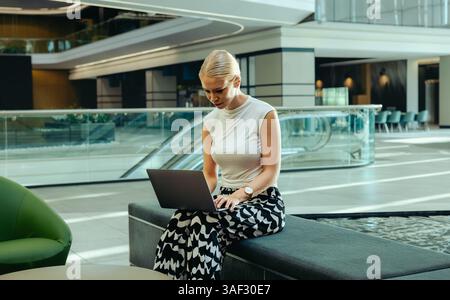A focused woman sits in a modern office lobby, engrossed in her work on a laptop. The open and contemporary environment conveys professionalism and mo Stock Photo