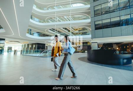 Two female corporate professionals walk and converse in a spacious, modern office lobby with sleek architectural design. The image captures the casual Stock Photo