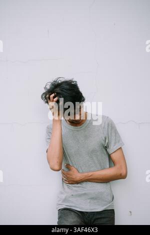 A young man displaying distress, scratching head against a textured wall. Demonstrates sadness, stress, anxiety, or frustration Stock Photo