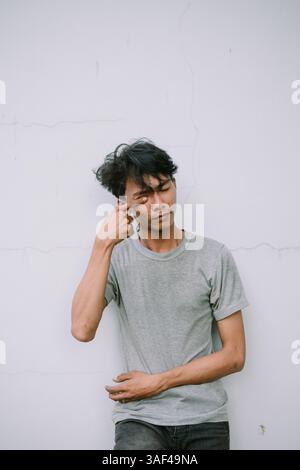 Pensive asian man touching his eye against a white textured backdrop. Close-up portrait conveys a sense of introspection Stock Photo