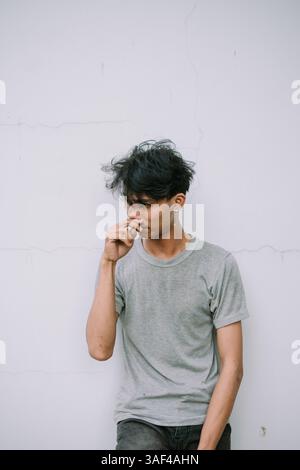 Contemplative young man in gray t-shirt leaning on a white wall. Minimalist portrait shows a modern, thoughtful mood. Stock Photo