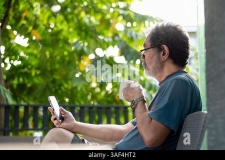 Senior Man using phone outdoors while drinking coffee at home Stock Photo
