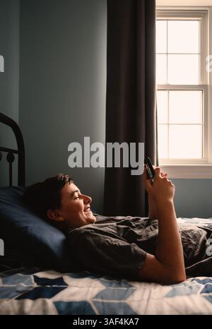 Teen boy in bedroom laughing using smartphone while laying on bed. Stock Photo