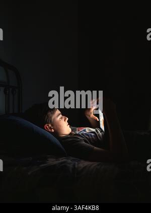 Teen boy in bedroom using tablet while laying on his bed at night. Stock Photo
