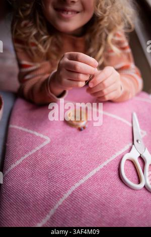 Child threading a needle while crafting on a pink textile surface Stock Photo