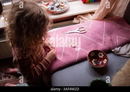 Child threading a needle while crafting on a pink textile surface Stock Photo