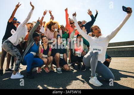 A diverse group of runners celebrates by taking a joyful selfie after completing a race. They're dressed in athletic wear with race numbers. Stock Photo