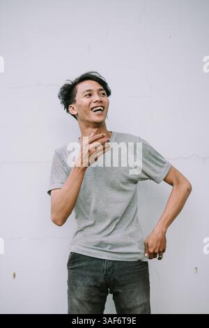 A happy young man wearing a gray shirt smiles broadly against a textured white wall with a joyful expression Stock Photo