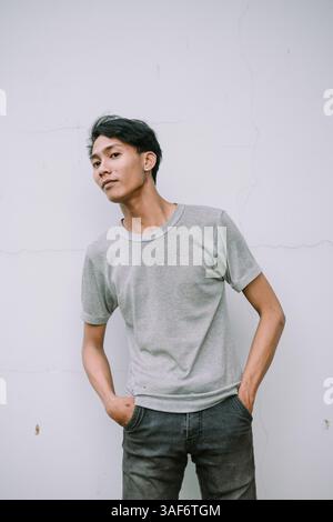 A young Asian man in casual wear confidently poses against a plain wall. His modern style is a blend of simplicity and urban trend. Stock Photo