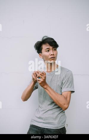 Pensive young man in casual wear stands against a plain wall in a thoughtful pose looking away. Stock Photo